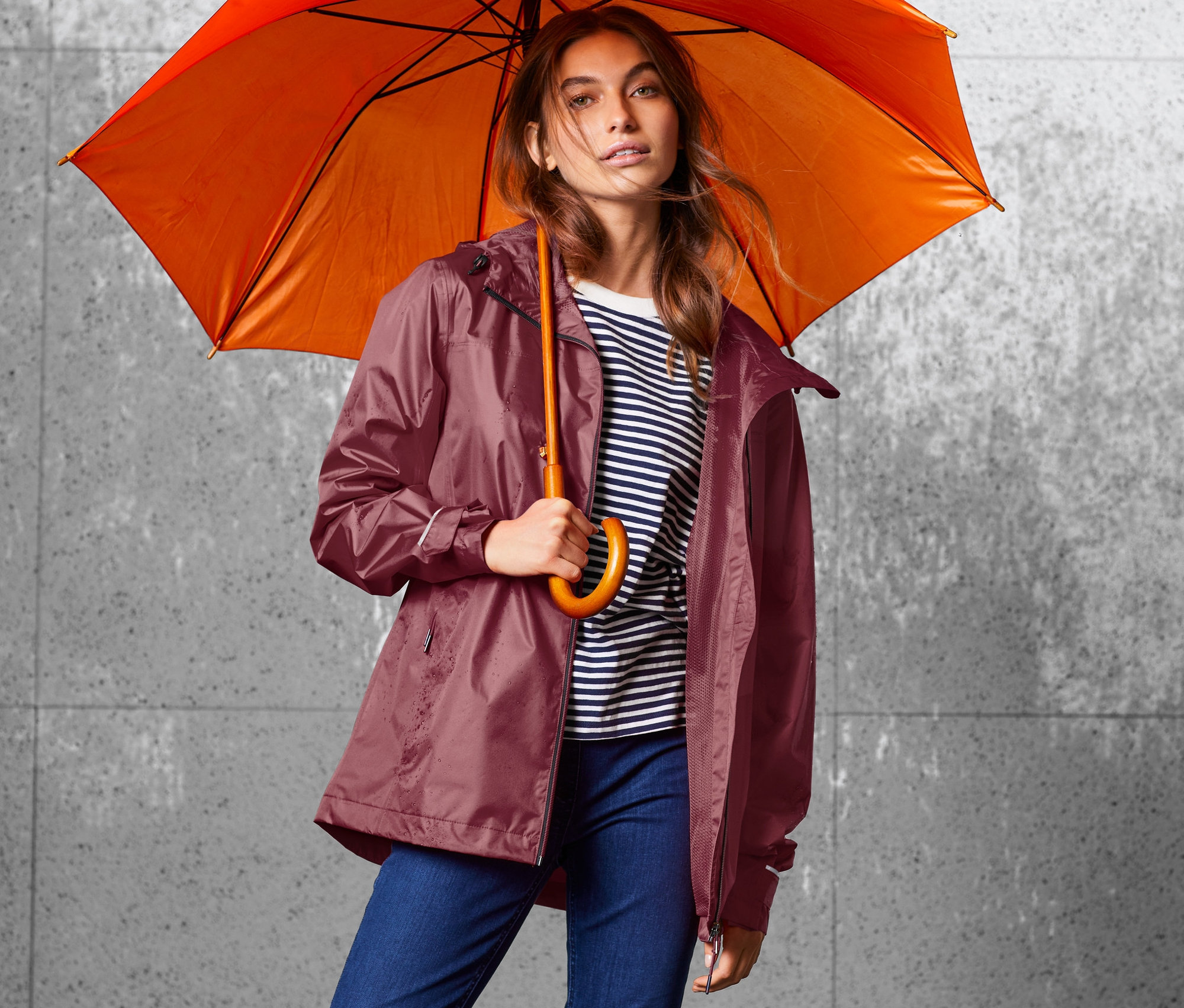 Femme tenant un parapluie orange et portant une veste de pluie bordeaux.