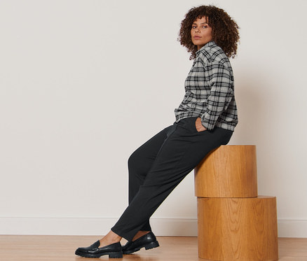 Femme assise sur des marches en bois portant un sweat-shirt à carreaux, un pantalon de jogging noir avec nervure et des loafers en cuir.