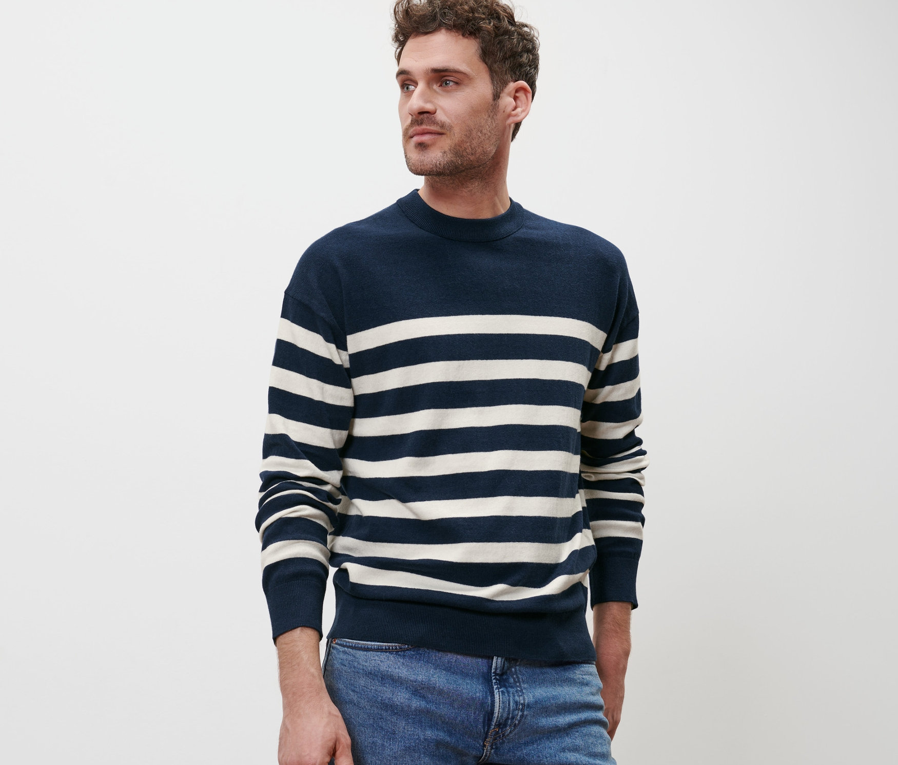 Un homme pose devant un fond blanc portant un pull à encolure ronde rayé bleu et blanc et un jean bleu.