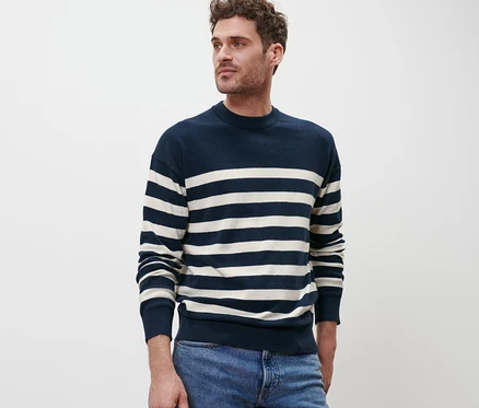 Un homme pose devant un fond blanc portant un pull à encolure ronde rayé bleu et blanc et un jean bleu.