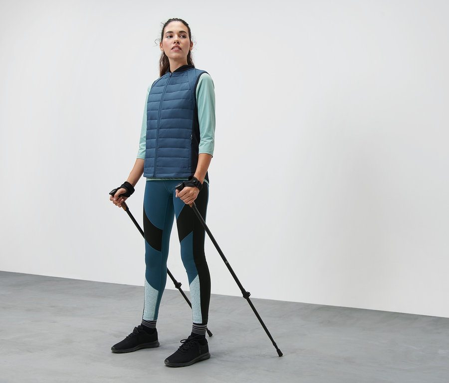 Femme tenant des bâtons de marche télescopiques. Elle porte un gilet matelassé thermique, un legging de sport color block et un t-shirt fonctionnel manches 3/4.