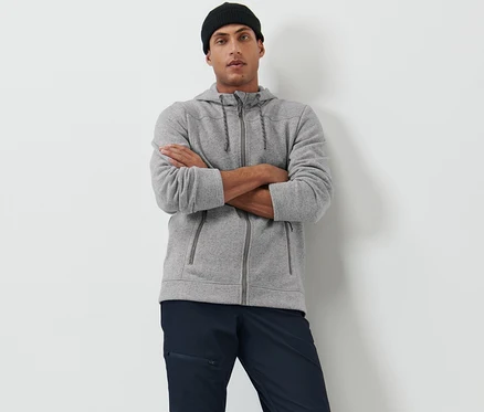 Un homme pose, les bras croisés, portant une veste en maille polaire grise et un bonnet noir.