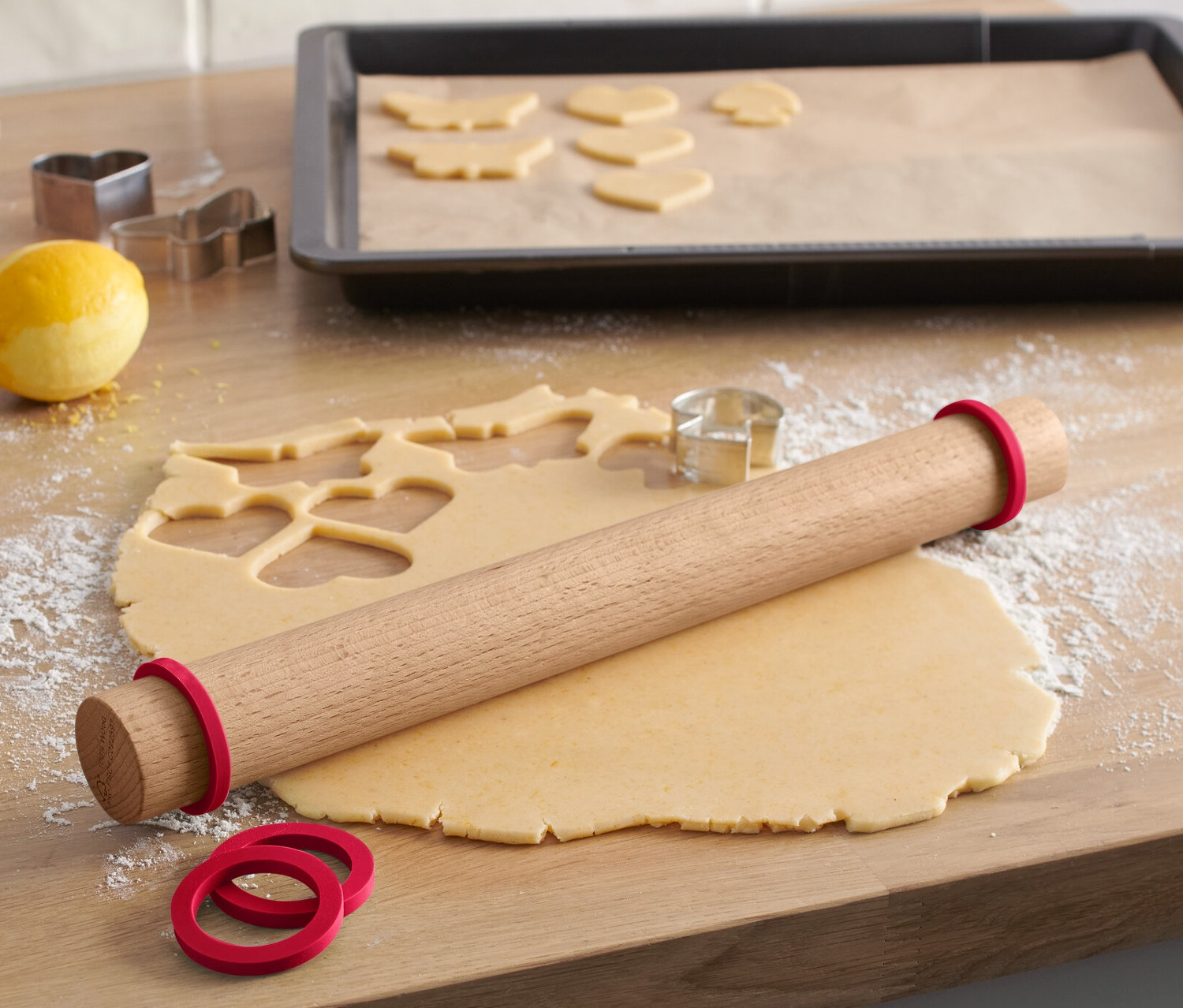 Sur une surface en bois reposent un rouleau à pâtisserie en bois avec des élastiques rouges, à côté de la pâte étalée avec des formes découpées et des emporte-pièces.