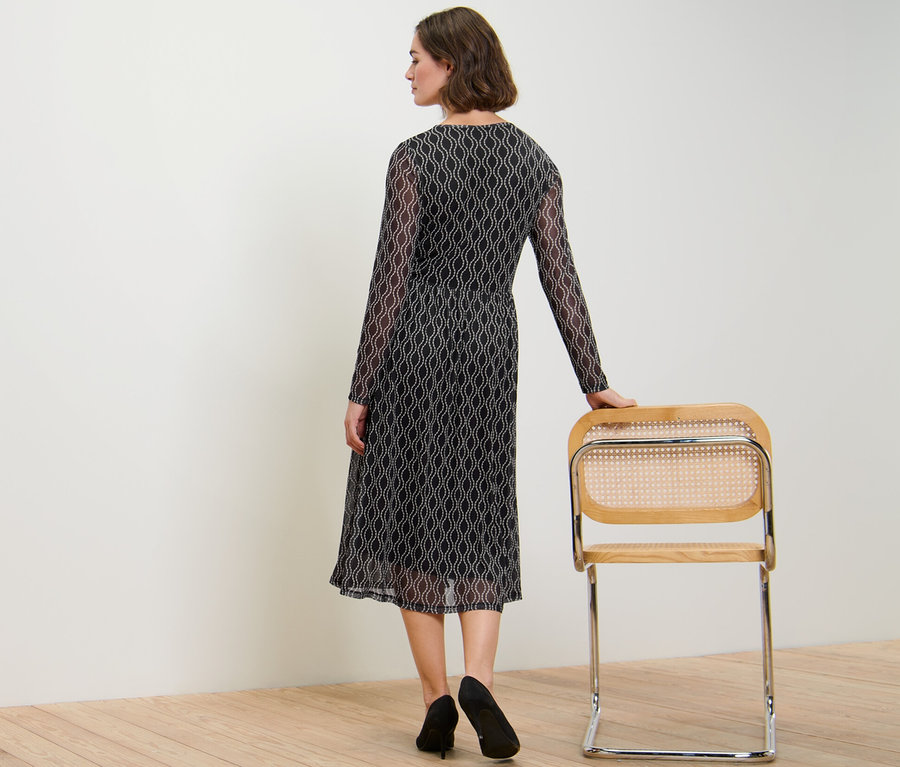 Femme vue de dos portant une robe en mesh imprimée, la main sur une chaise.