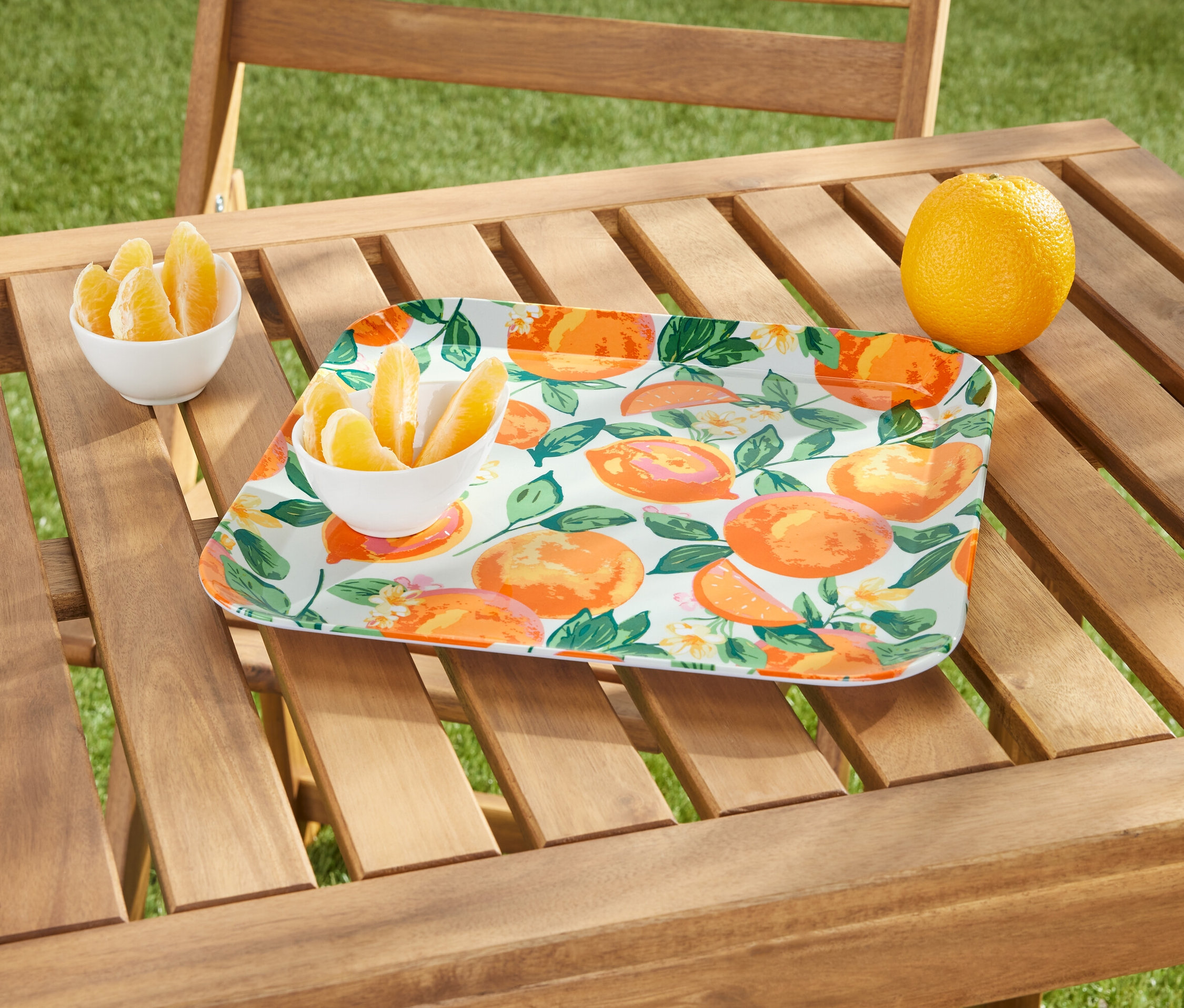 Sur une table en bois se trouvent un plateau à motif d'oranges, un bol de quartiers d'orange et une orange entière.