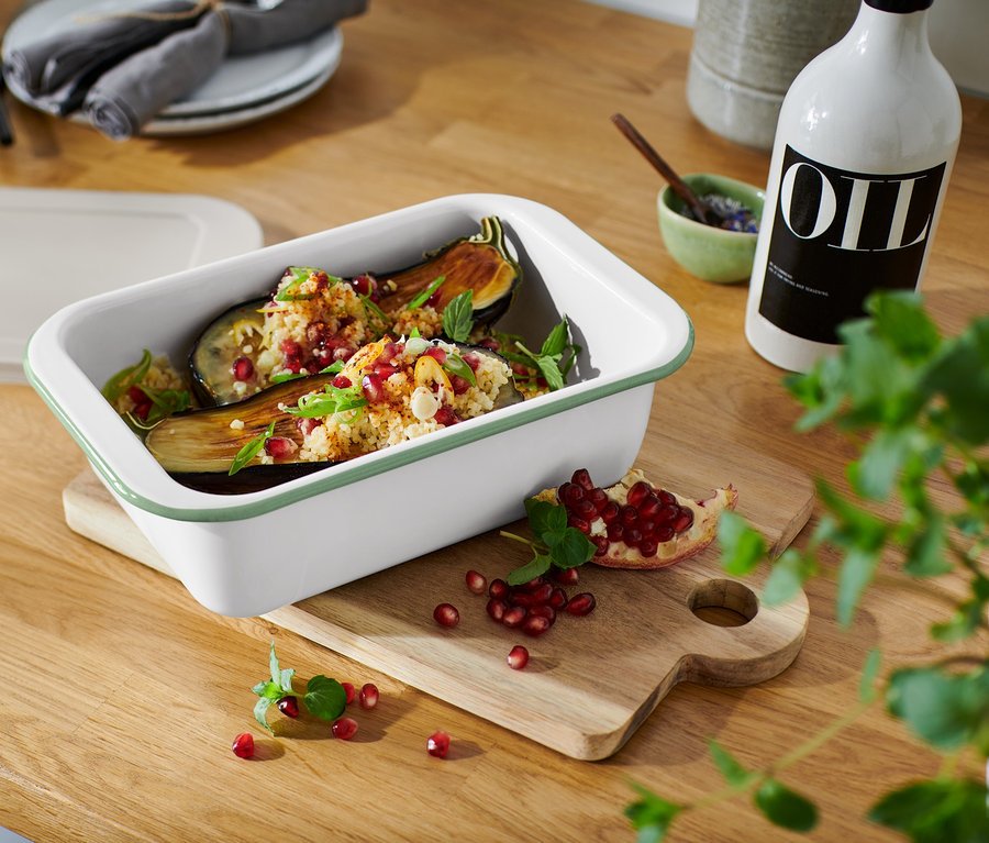 Aubergines cuites au four dans un petit moule à gratin en émail avec couvercle, parsemées de grenade, sur une planche à découper en bois.