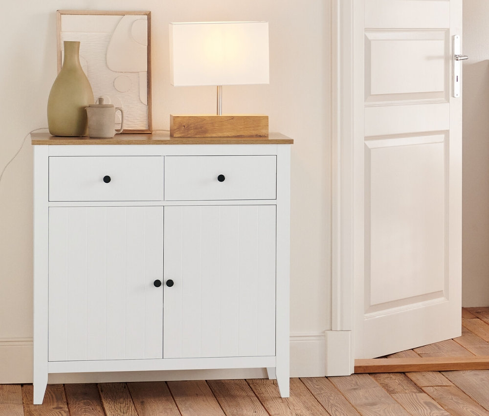 Dans la pièce, il y a une commode à tiroirs, un buffet, une étagère et une lampe de table avec un pied en bois.