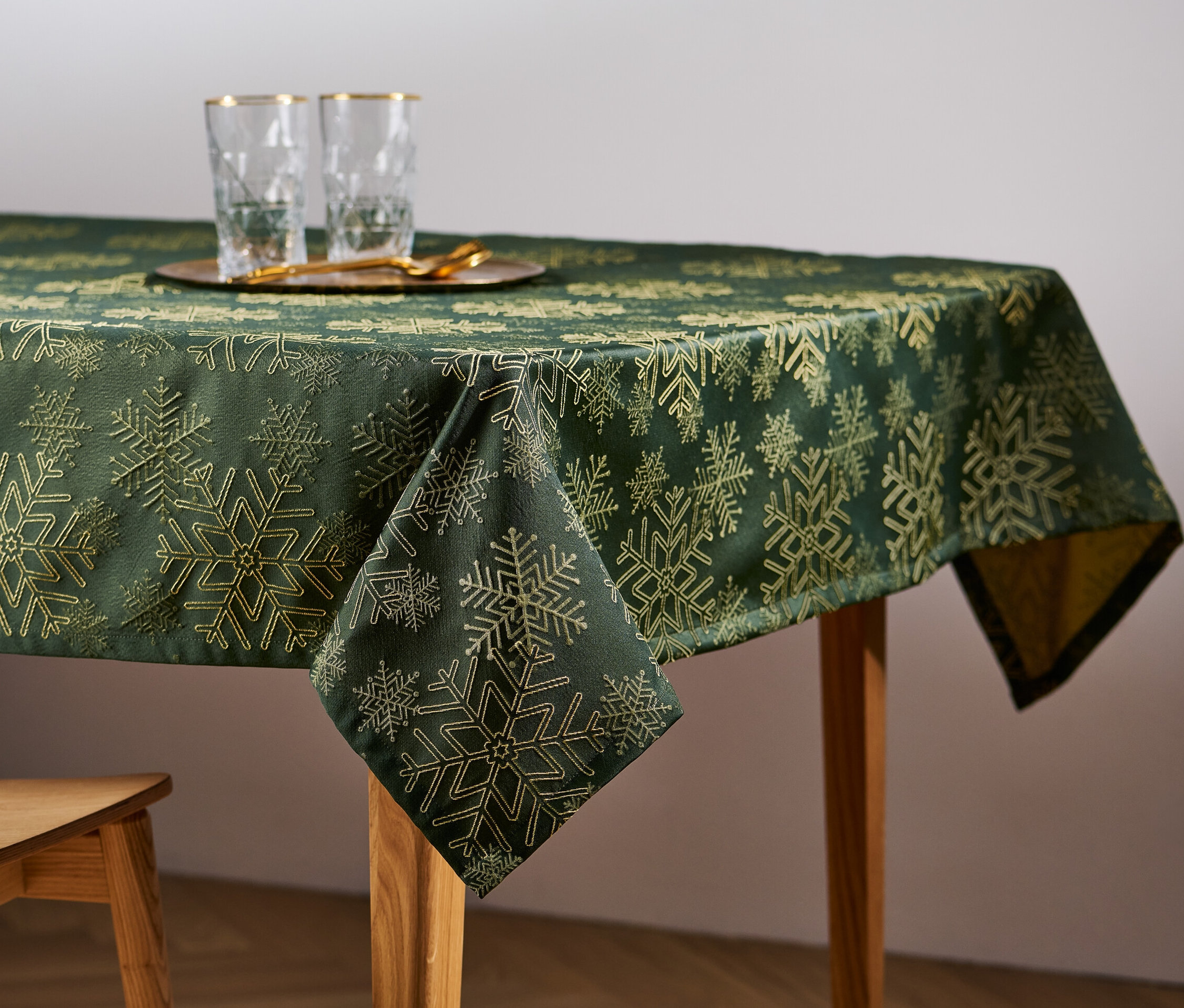 Sur une table en bois, une nappe jacquard verte avec un motif de flocons de neige. Sur la table, des verres et une chaise en bois.