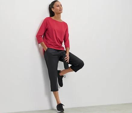 Une femme appuyée contre un mur portant un t-shirt rouge et un pantalon de sport 3/4.