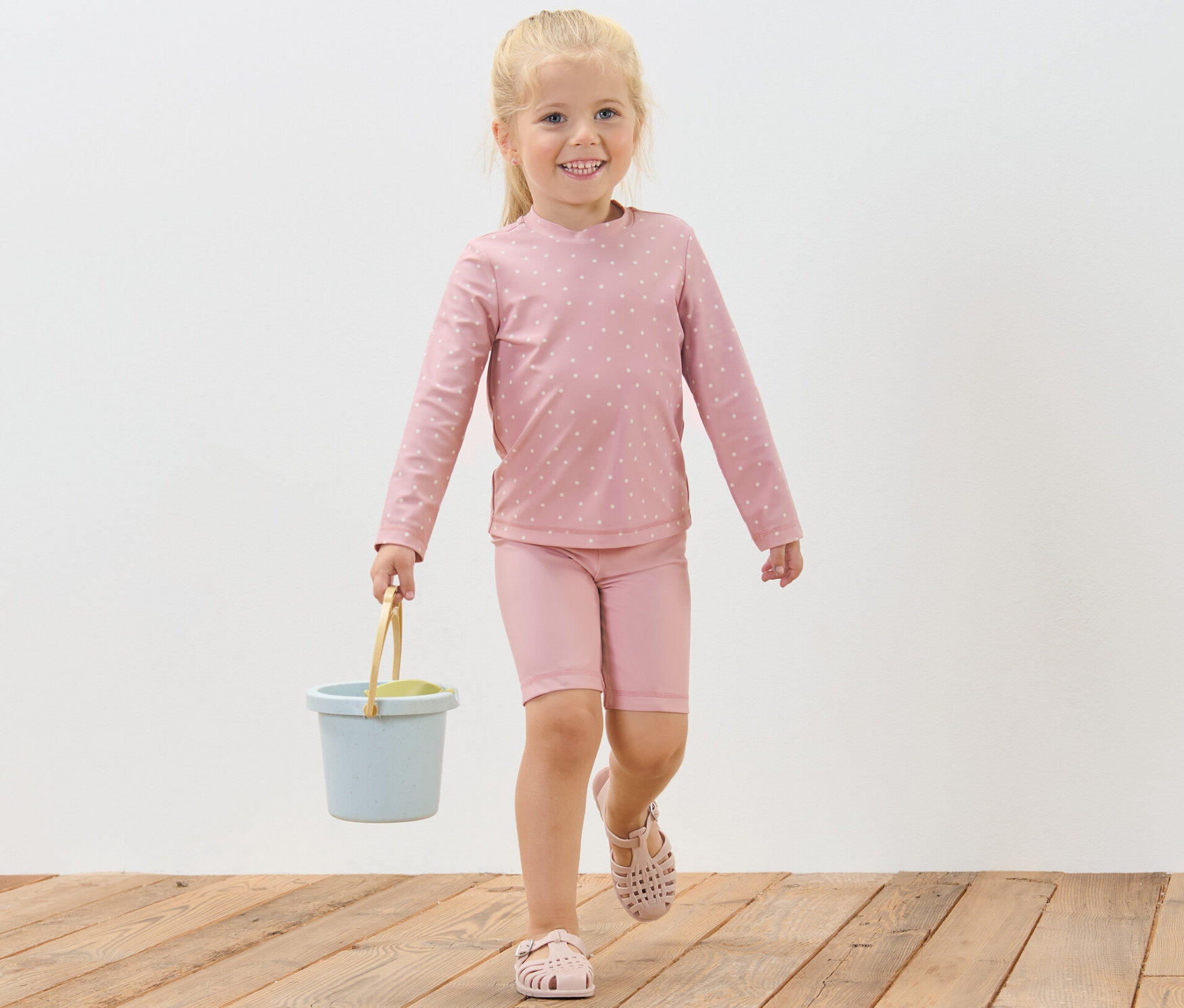 Une fillette souriante marche sur un plancher en bois et porte un seau bleu. Elle porte un ensemble pour enfant avec indice de protection UV 80, à pois.
