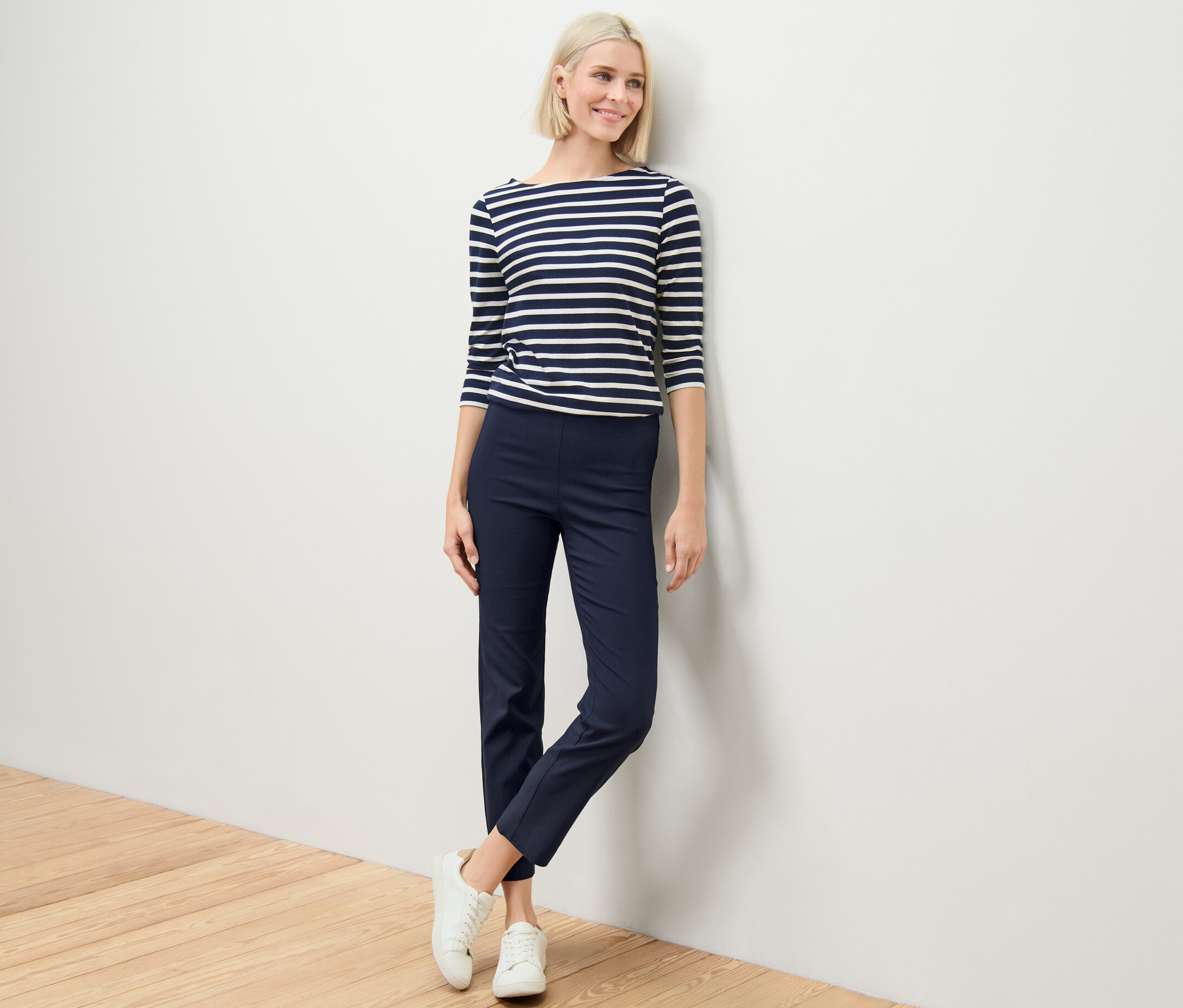 Une femme blonde se tient contre un mur et porte un pantalon stretch bleu marine.