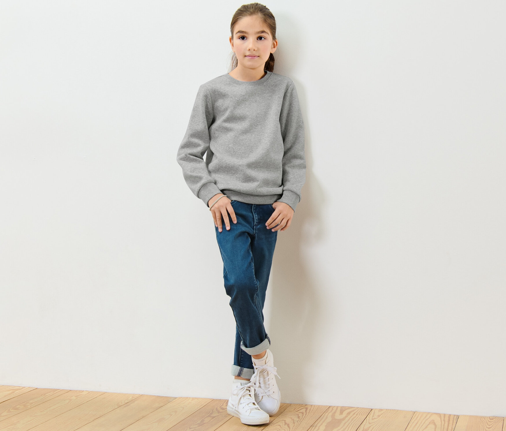 Une fille est appuyée contre un mur blanc et porte un sweat-shirt gris pour enfant avec un intérieur gratté.