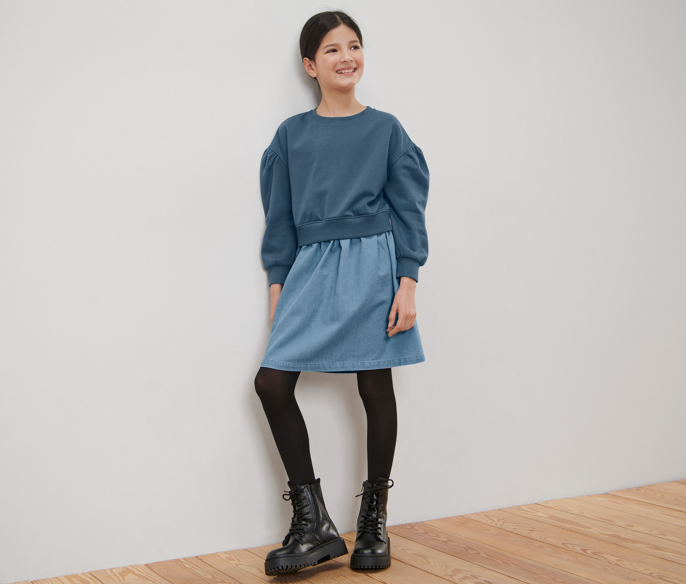 Une jeune fille souriante en robe en sweat pour enfant est appuyée contre un mur blanc.