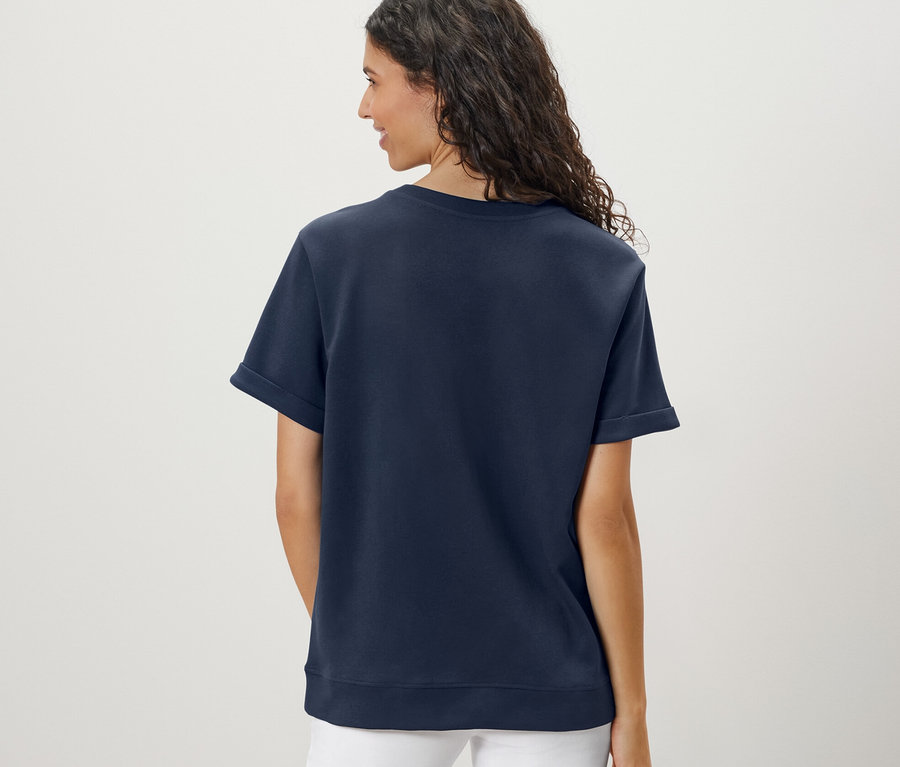 Vue arrière d'une femme aux cheveux ondulés portant un t-shirt bleu foncé en mélange modal et un pantalon blanc.