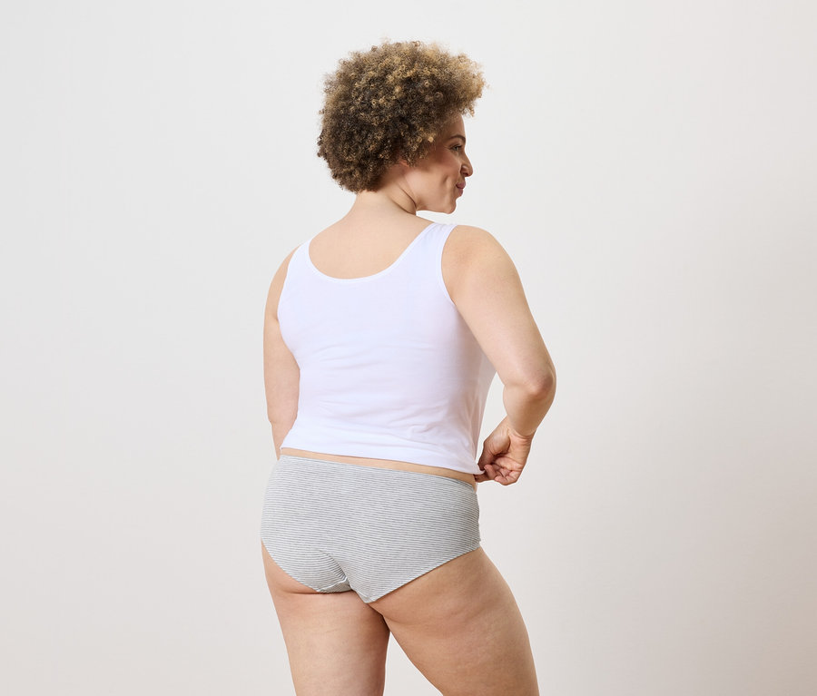 Une femme aux cheveux bouclés, portant un débardeur et une culotte rayée, se tient dos à nous.