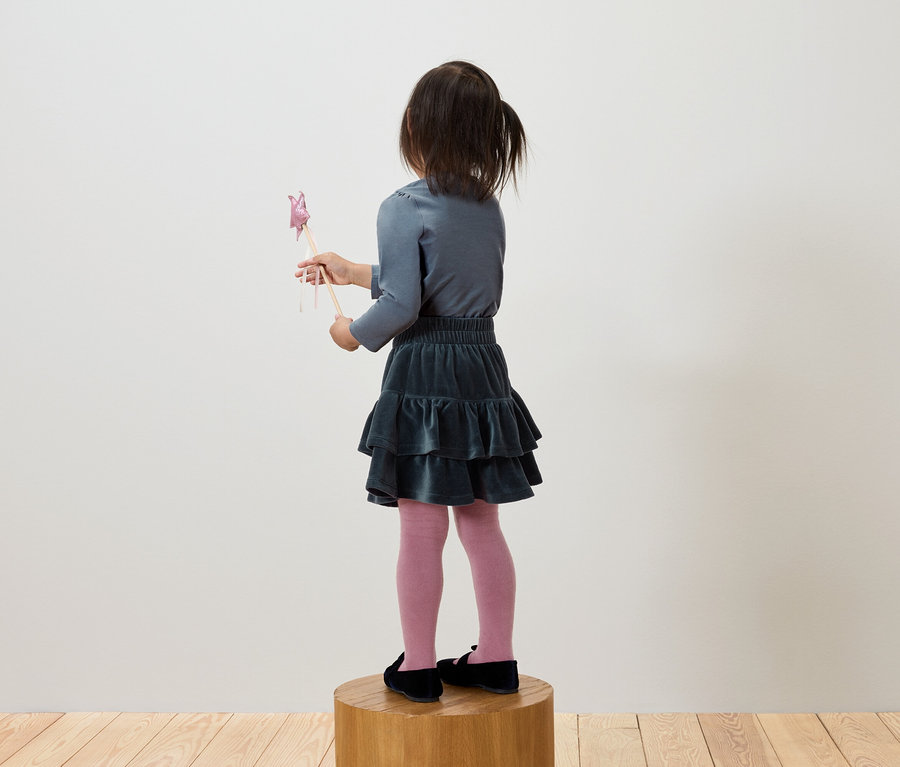 Une fille en t-shirt gris et jupe verte se tient sur un socle en bois et tient une baguette.