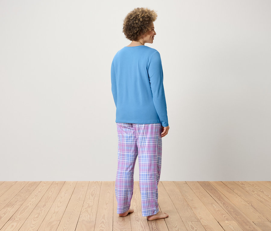 Une femme aux cheveux bouclés se tient dos à la caméra, portant un t-shirt bleu à manches longues et un pantalon à carreaux.