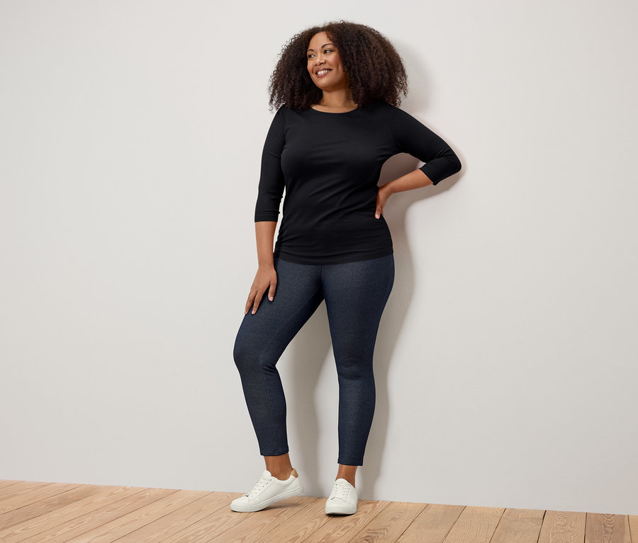 Femme souriante posant dans un t-shirt noir à manches trois-quarts, un jegging bleu foncé et des baskets blanches.
