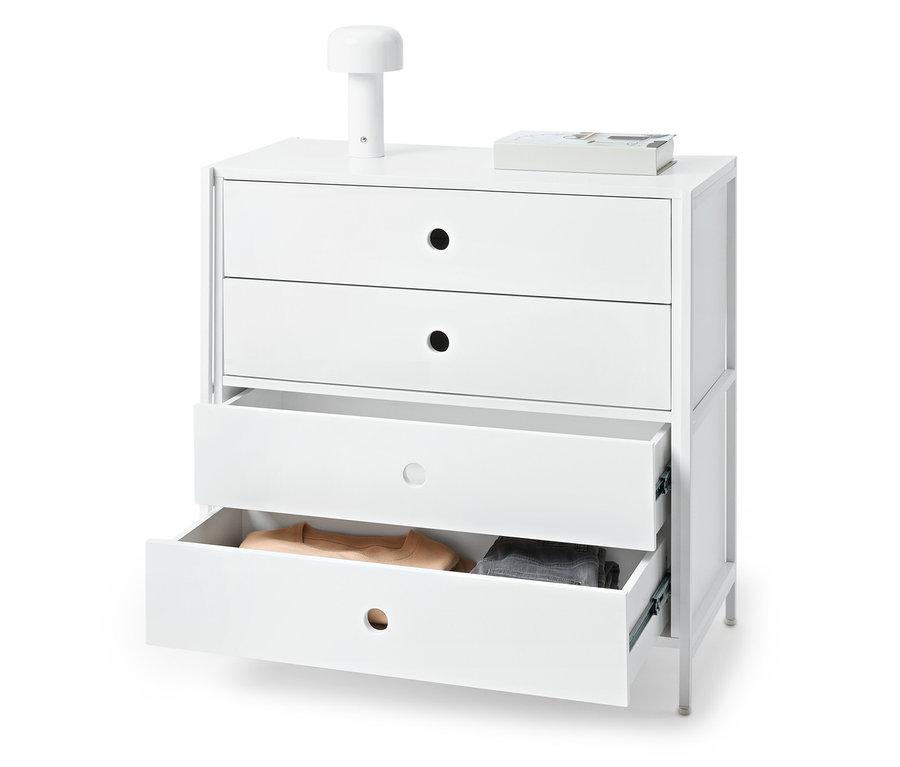 Commode blanche avec trois tiroirs dont deux ouverts et contenant des vêtements. Une lampe et un livre sont posés sur la commode.