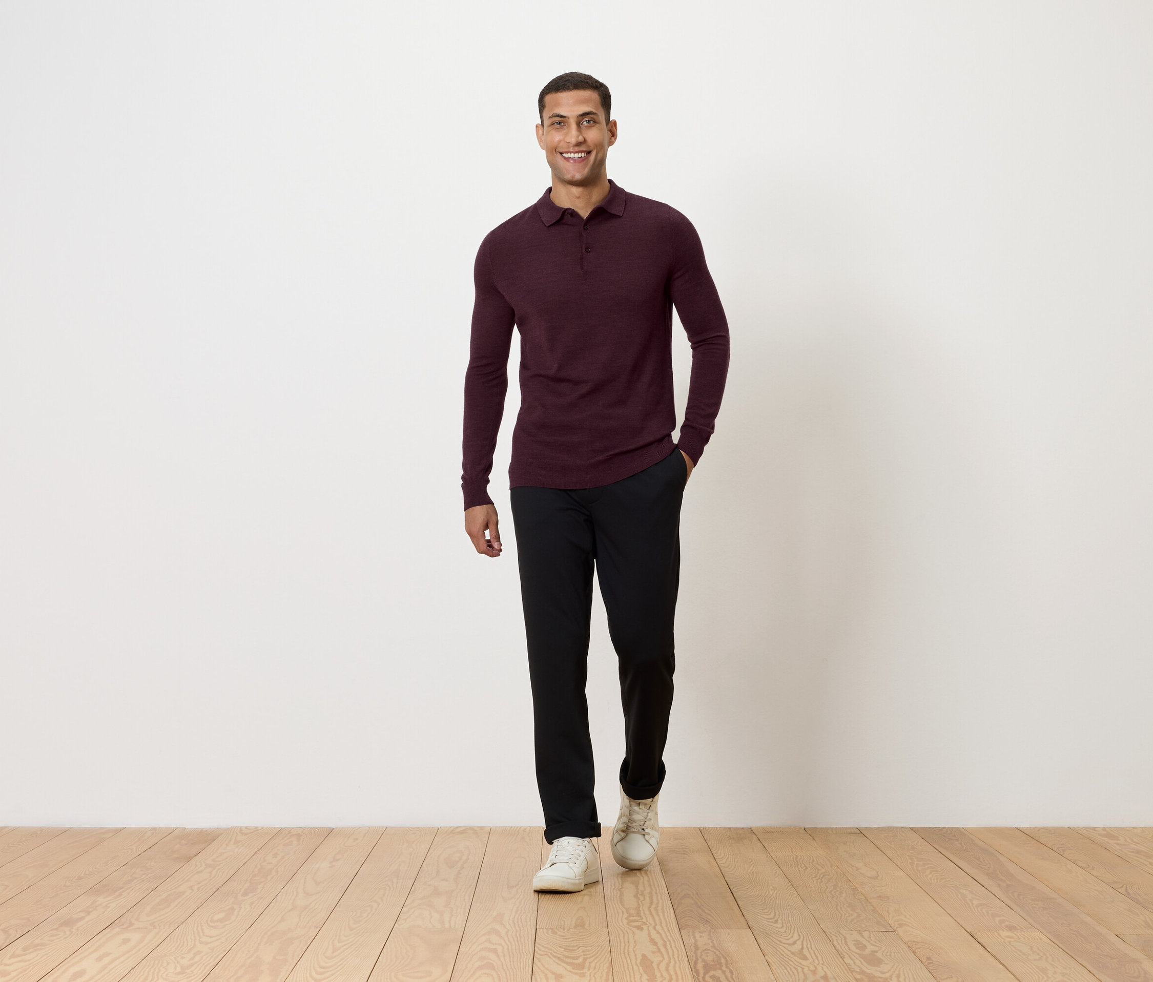 Un homme marche sur un plancher en bois. Il porte un polo bordeaux à manches longues, un pantalon noir et des baskets blanches.