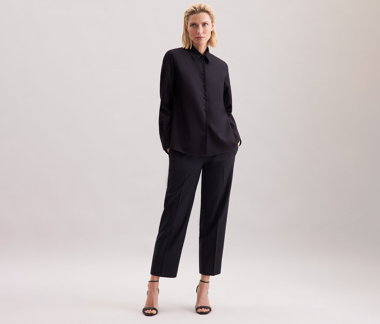 Une femme blonde se tient sur un fond clair en chemisier noir Seidensticker, un pantalon noir et des sandales noires.