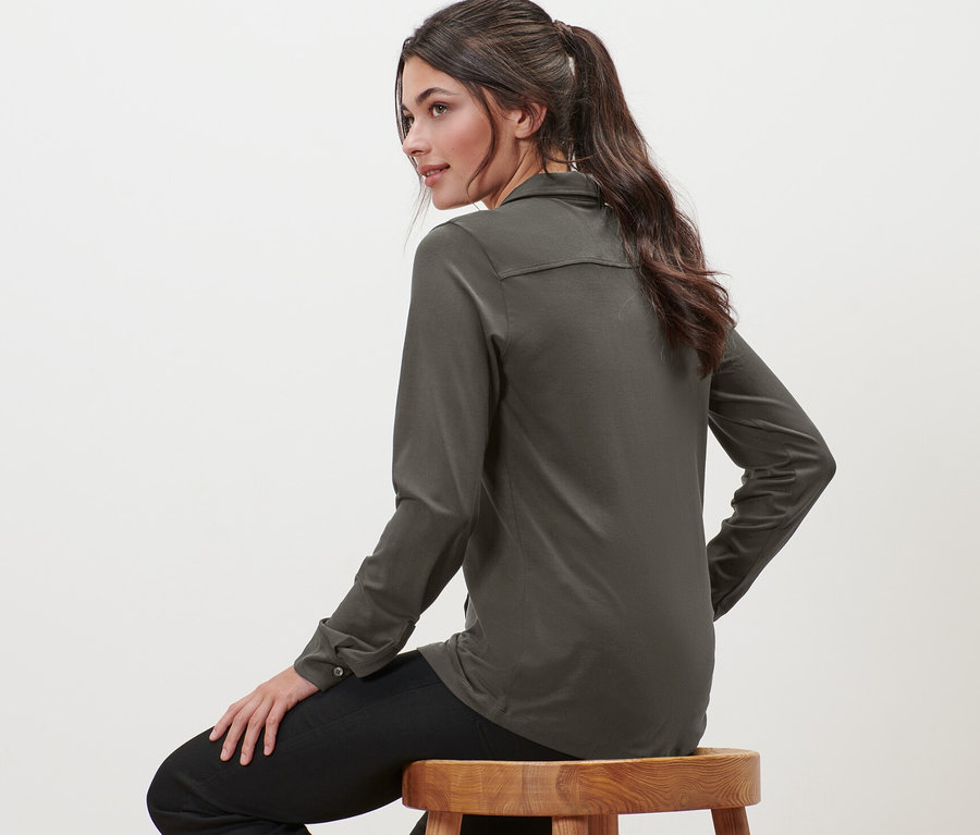 Femme assise sur un tabouret en bois portant une blouse avec patte de boutonnage.