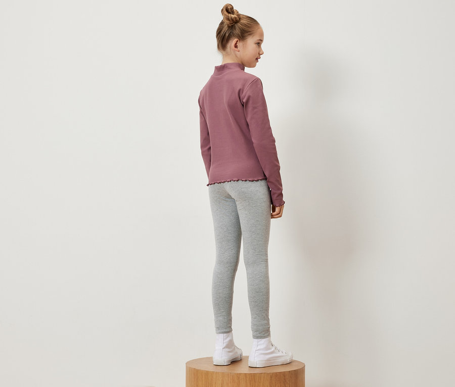 Une fille se tient de côté sur un socle en bois, portant un t-shirt à manches longues rose, un legging gris et des baskets blanches.