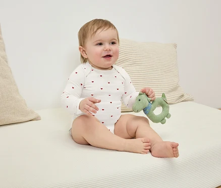 Un bébé assis portant un body à motifs de cœurs rouges tenant un jouet dinosaure vert.