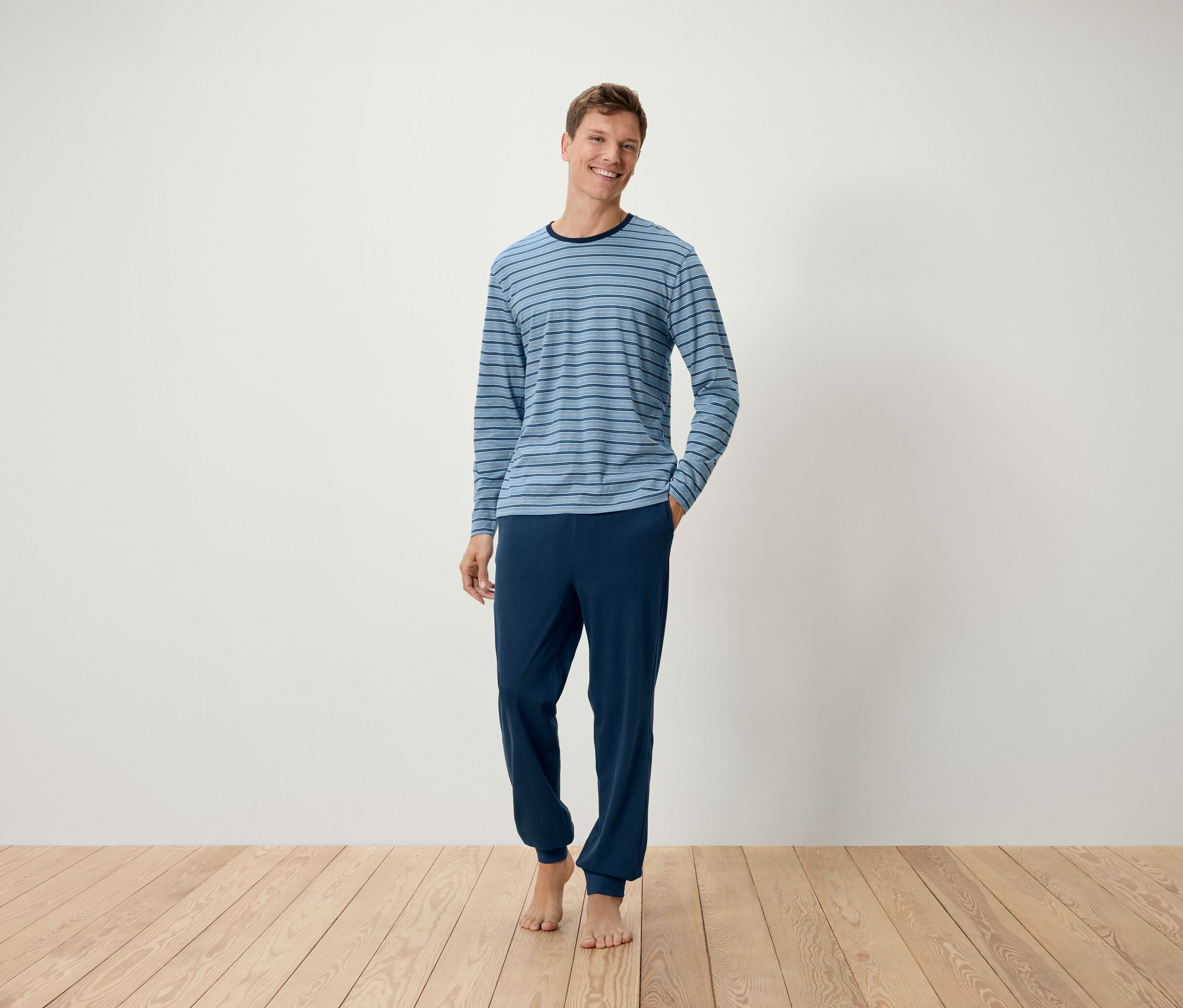 Un homme se tient pieds nus sur un plancher en bois, portant un pyjama à ourlets bleu clair à rayures.