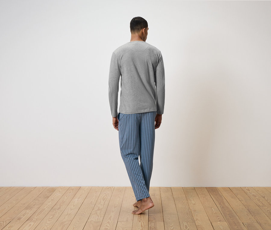 Un homme, dos à la caméra, se tient sur un plancher en bois. Il porte un t-shirt gris à manches longues et un pantalon de pyjama rayé bleu.