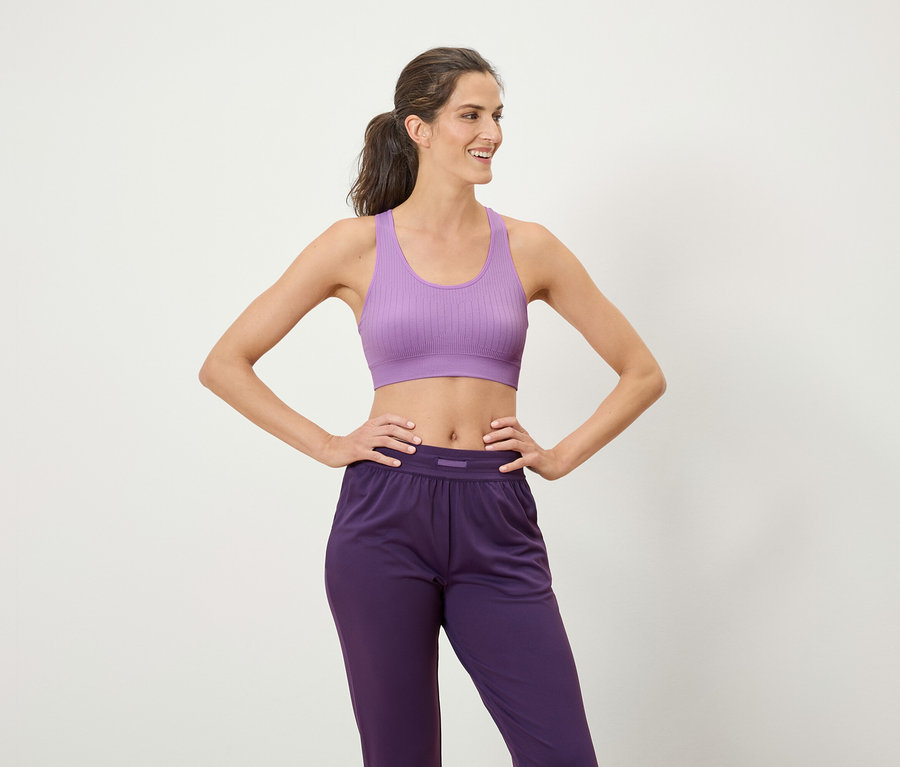 Femme brune aux cheveuxQueue de cheval posant dans un soutien-gorge de sport lilas et un pantalon de sport violet.