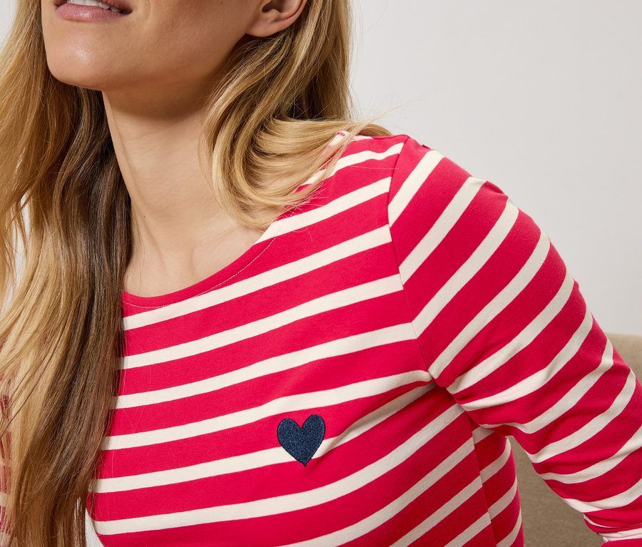 Une femme blonde porte un t-shirt à manches longues rayé rouge et blanc avec un cœur bleu marine.