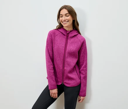 Une femme souriante pose en sweat à capuche rose et pantalon noir.