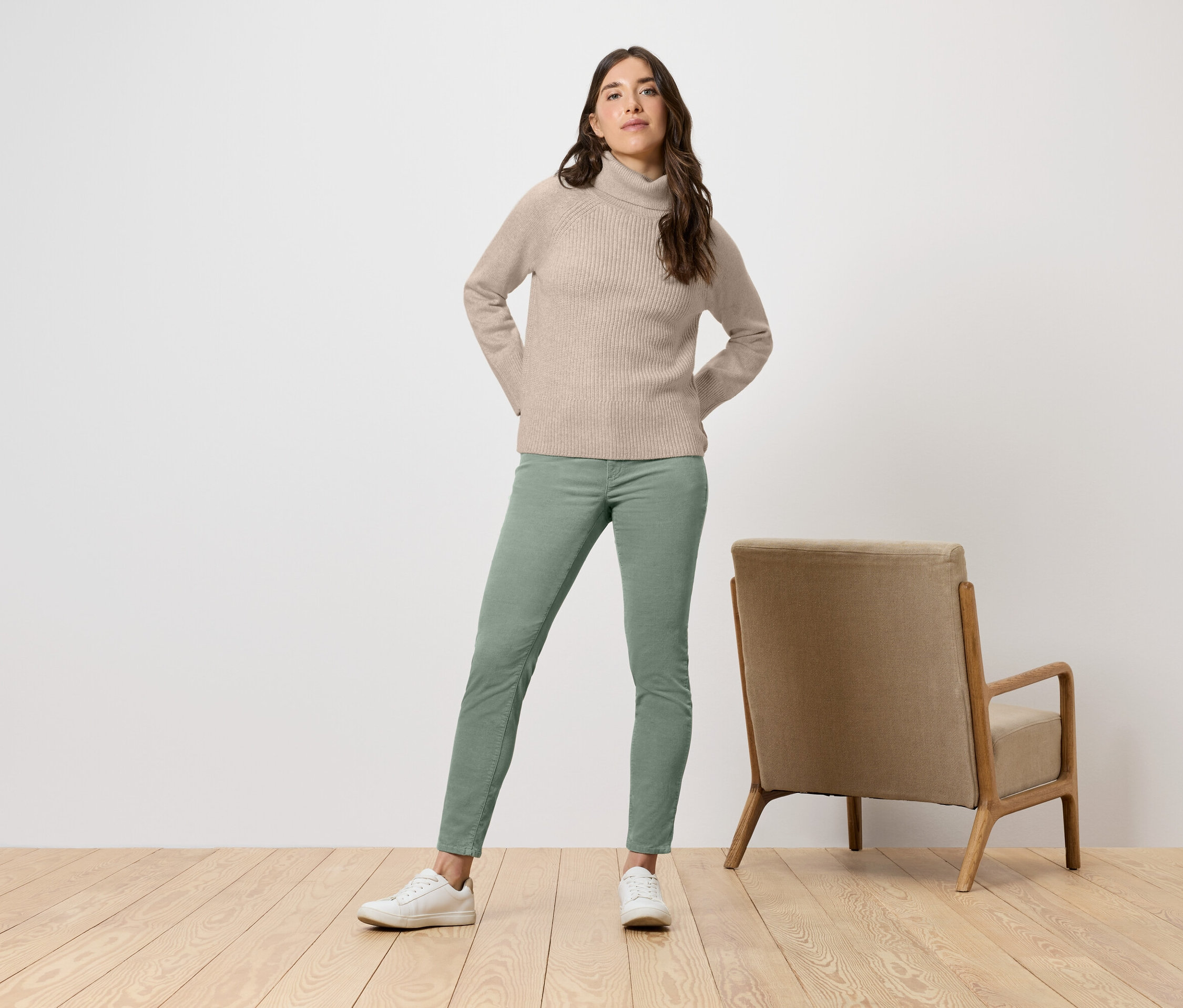 Femme en col roulé beige et pantalon vert debout à côté d'un fauteuil beige.