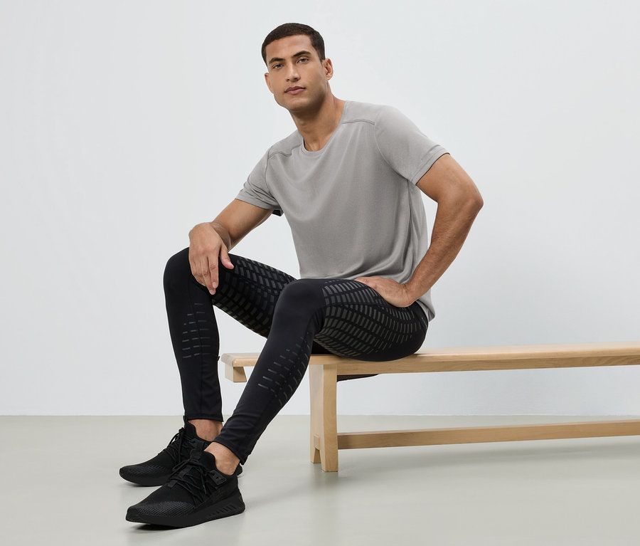 Un homme est assis sur un banc, portant un t-shirt gris, un legging noir à motifs et des baskets noires.