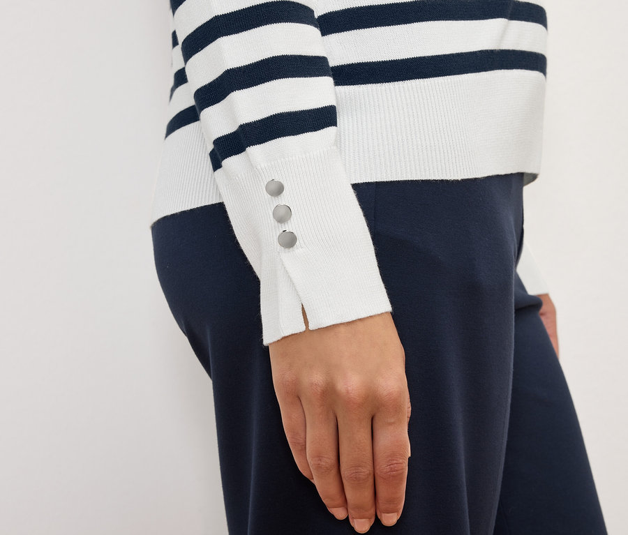 Une femme porte un pull rayé à manches longues avec des boutons aux poignets et un pantalon bleu marine.