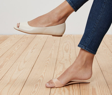 Jambes d'une femme en jean, une jambe portant une ballerine crème à bout ouvert et l'autre une sandale à lanières. Une paire de semelles intérieures rembourrées est dans la sandale.