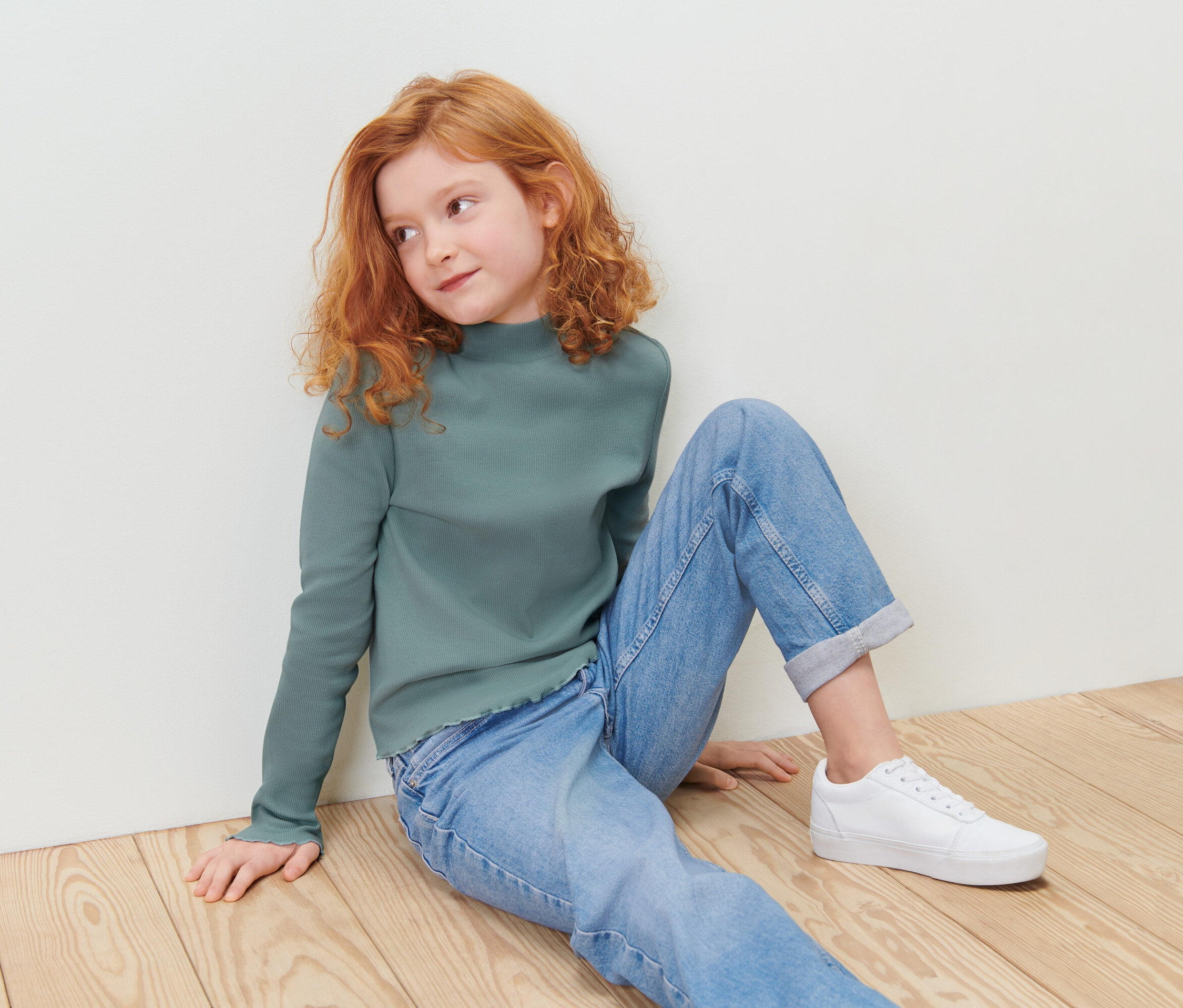 Une fille rousse est assise sur un plancher en bois, portant un t-shirt vert à manches longues et un jean bleu.
