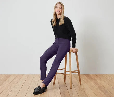 Une femme blonde est assise sur un tabouret en bois, portant un haut noir et un pantalon violet.