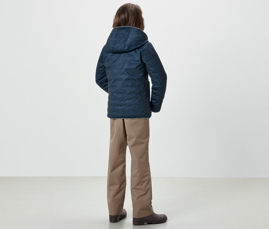 Un garçon se tient dos à la caméra portant une veste bleue et un pantalon de pluie pour enfants.