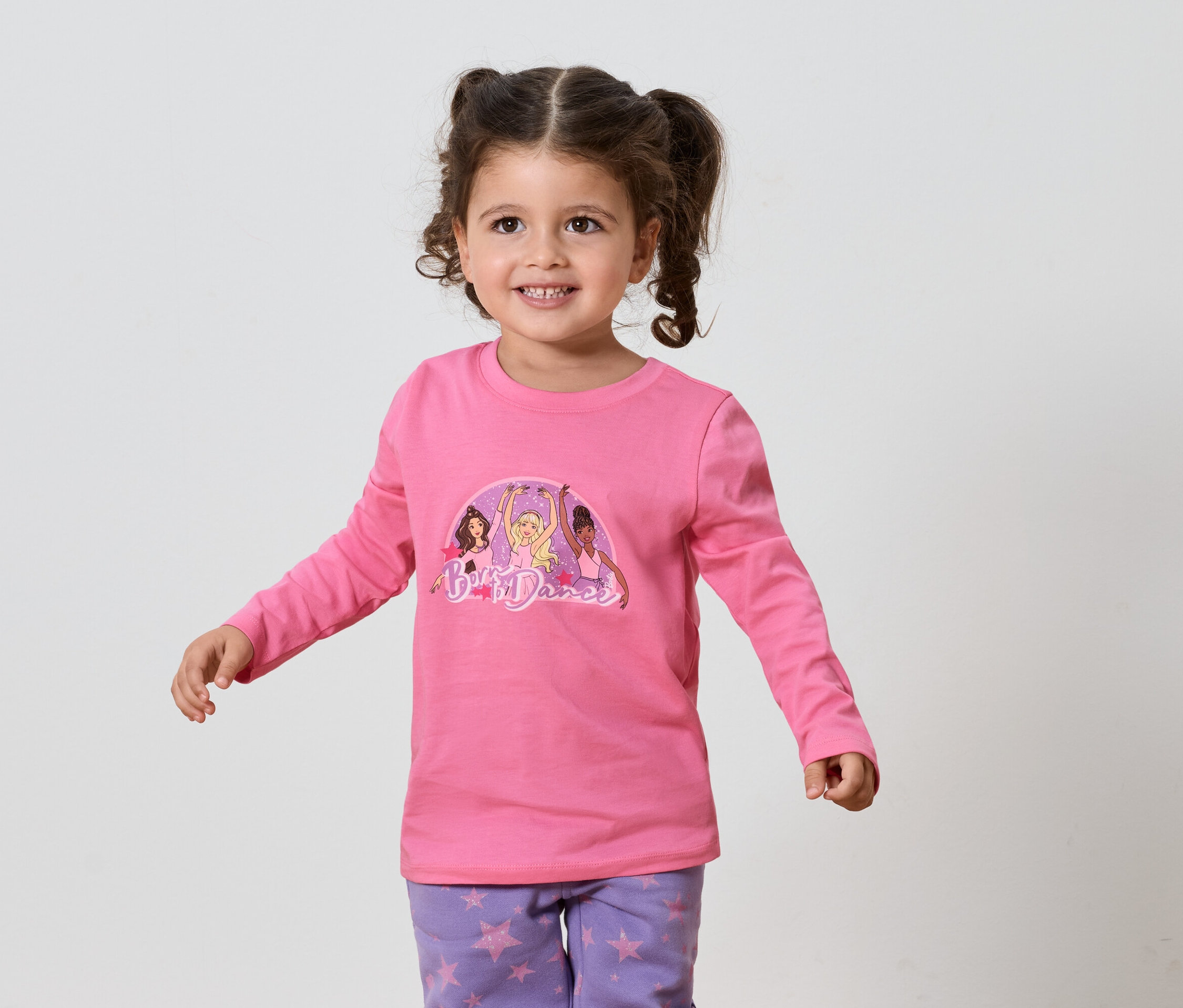 Petite fille souriante aux cheveux bruns, portant un t-shirt rose à manches longues et un pantalon violet à motifs d'étoiles.