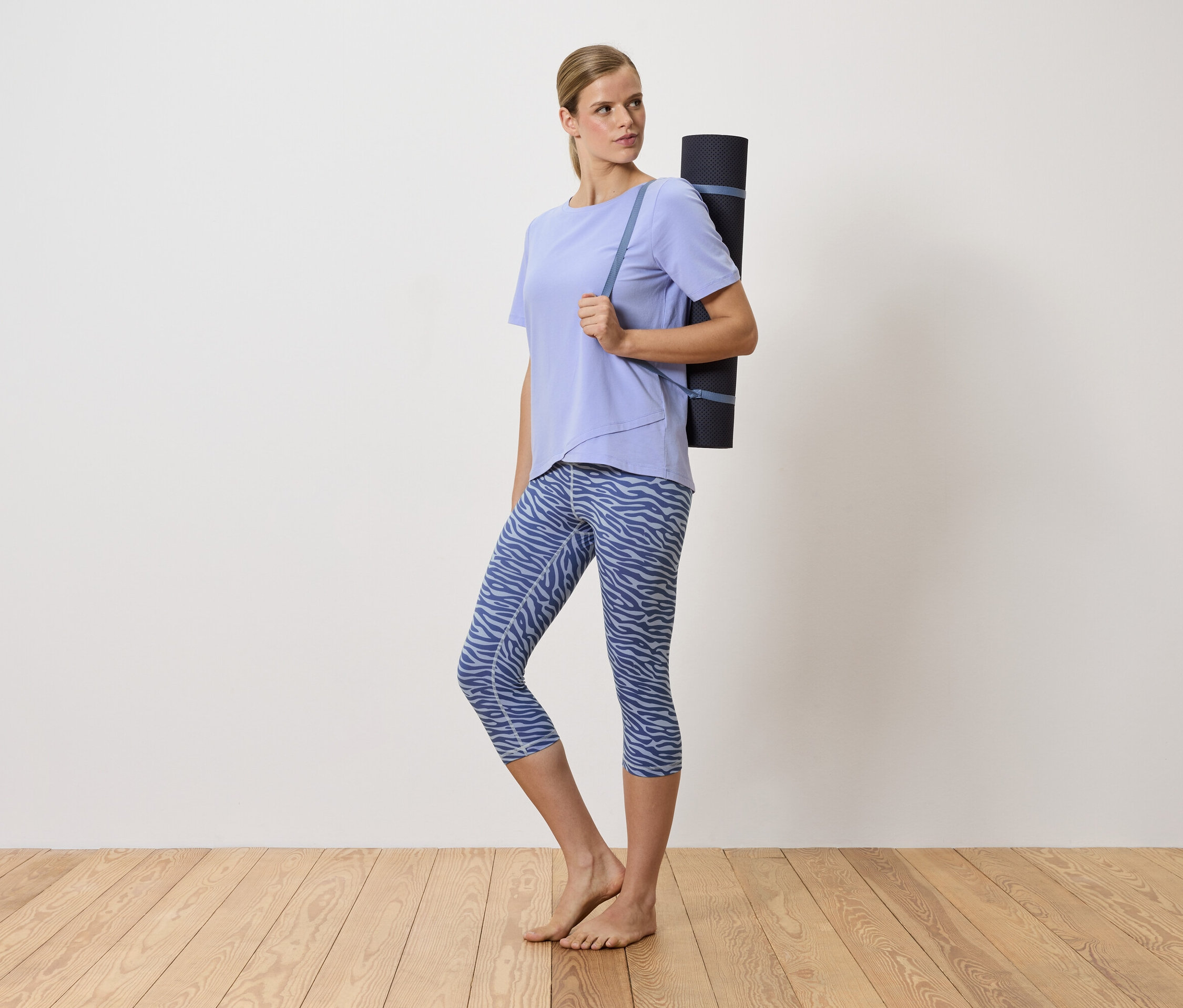 Une femme blonde porte un tapis de yoga sur l'épaule. Elle porte un t-shirt violet et un legging bleu à motifs.