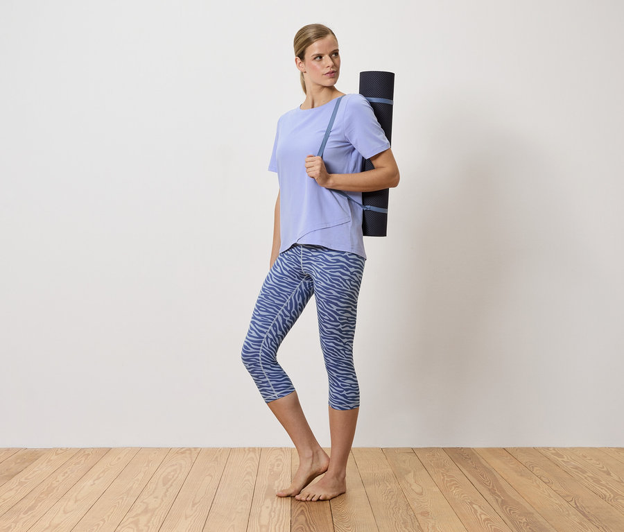 Une femme blonde porte un tapis de yoga sur l'épaule. Elle porte un t-shirt violet et un legging bleu à motifs.