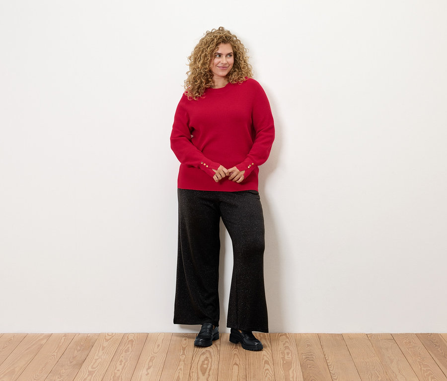 Une femme aux cheveux bouclés se tient debout et porte un pull rouge et un pantalon noir.