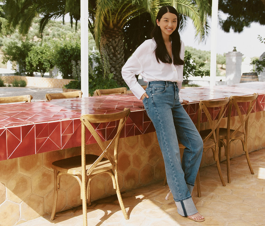 Une femme pose, appuyée sur un bar à l'extérieur, portant un jean Angels « Lara » coupe droite avec revers bleu clair.