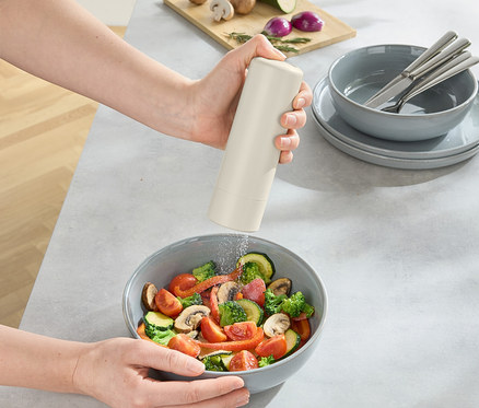 Une personne assaisonne un saladier, une planche à découper avec des champignons, des oignons et des courgettes est posée sur la table.