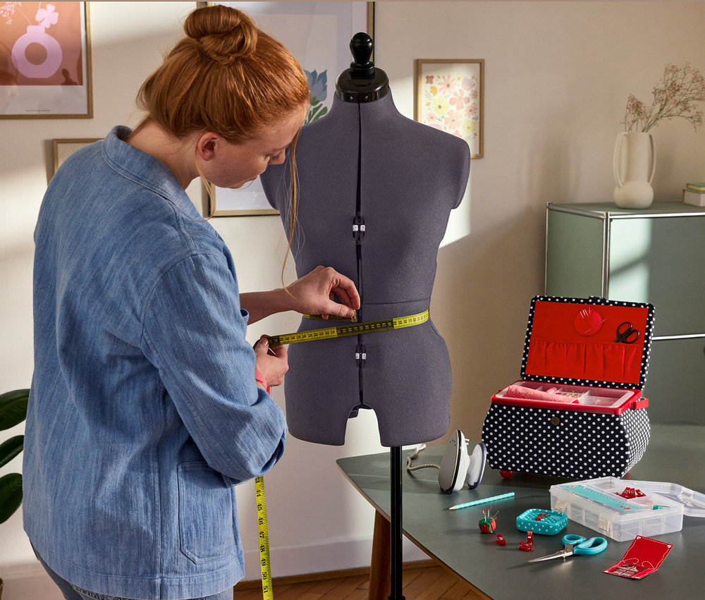 Une femme mesure la taille sur le buste de couture Prym Multi-Schneiderbüste »Prymadonna«. À côté : Prym Nähkorb »Polka Dots«, Prym Mini-Dampfbügeleisen, Prym Starter-Set Nähen »Love«.