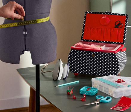 Une femme mesure la taille sur le buste de couture Prym Multi-Schneiderbüste »Prymadonna«. À côté : Prym Nähkorb »Polka Dots«, Prym Mini-Dampfbügeleisen, Prym Starter-Set Nähen »Love«.