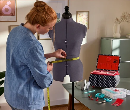 Une femme mesure la taille sur le buste de couture Prym Multi-Schneiderbüste »Prymadonna«. À côté : Prym Nähkorb »Polka Dots«, Prym Mini-Dampfbügeleisen, Prym Starter-Set Nähen »Love«.