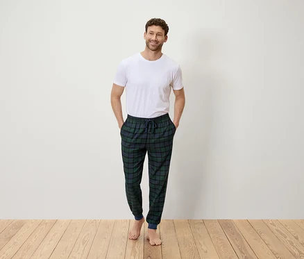 Un homme se tient debout, les mains dans les poches, portant un t-shirt blanc et un pantalon à carreaux vert et bleu.